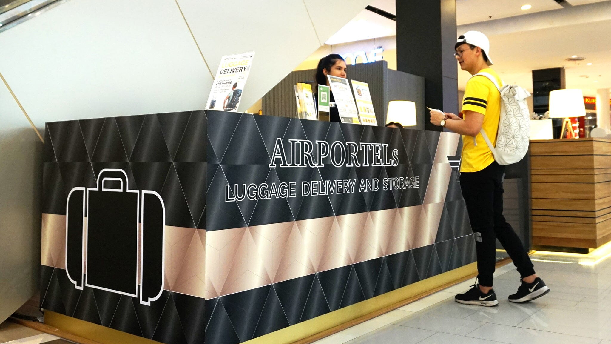 A traveller checking a bag in at a staffed AIRPORTELs counter inside a Bangkok shopping mall, with a member of our own team behind the desk