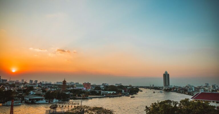 Photo: golden-hour traveler leaning on a Chao Phraya riverside railing with only a small daypack, Wat Arun glowing acros