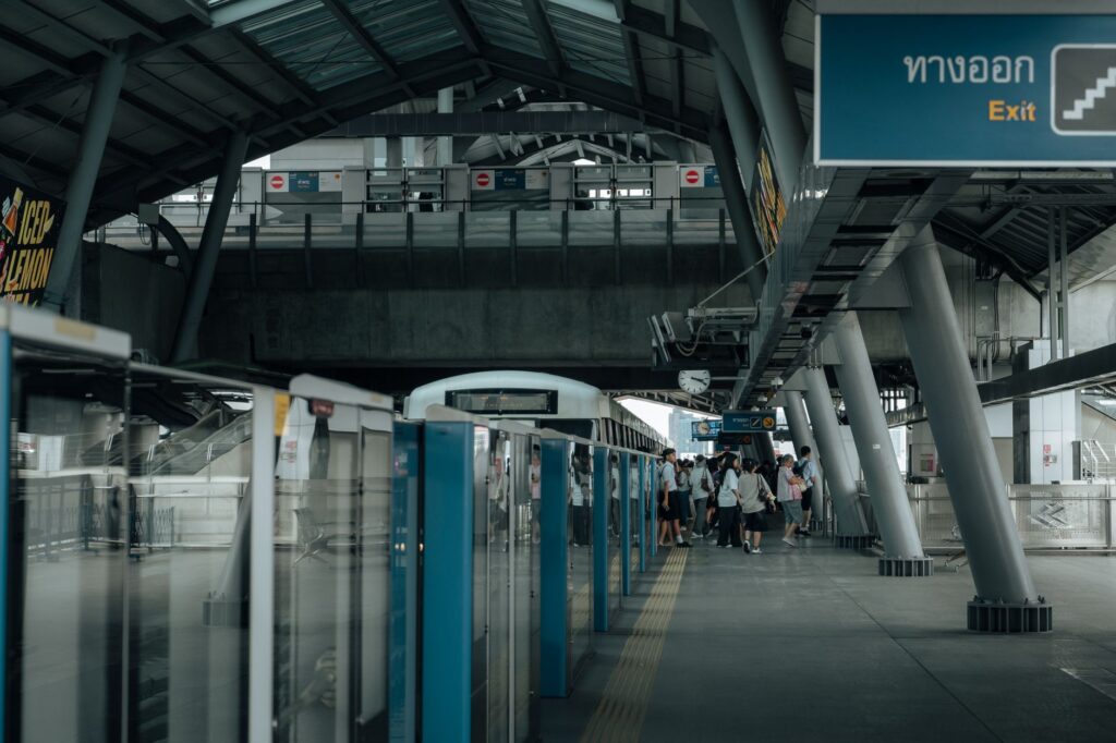 A Bangkok skytrain platform with passengers waiting behind platform screen doors, under a Thai and English exit sign