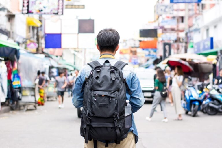A backpacker seen from behind, walking down a busy street of shops, stalls and parked scooters
